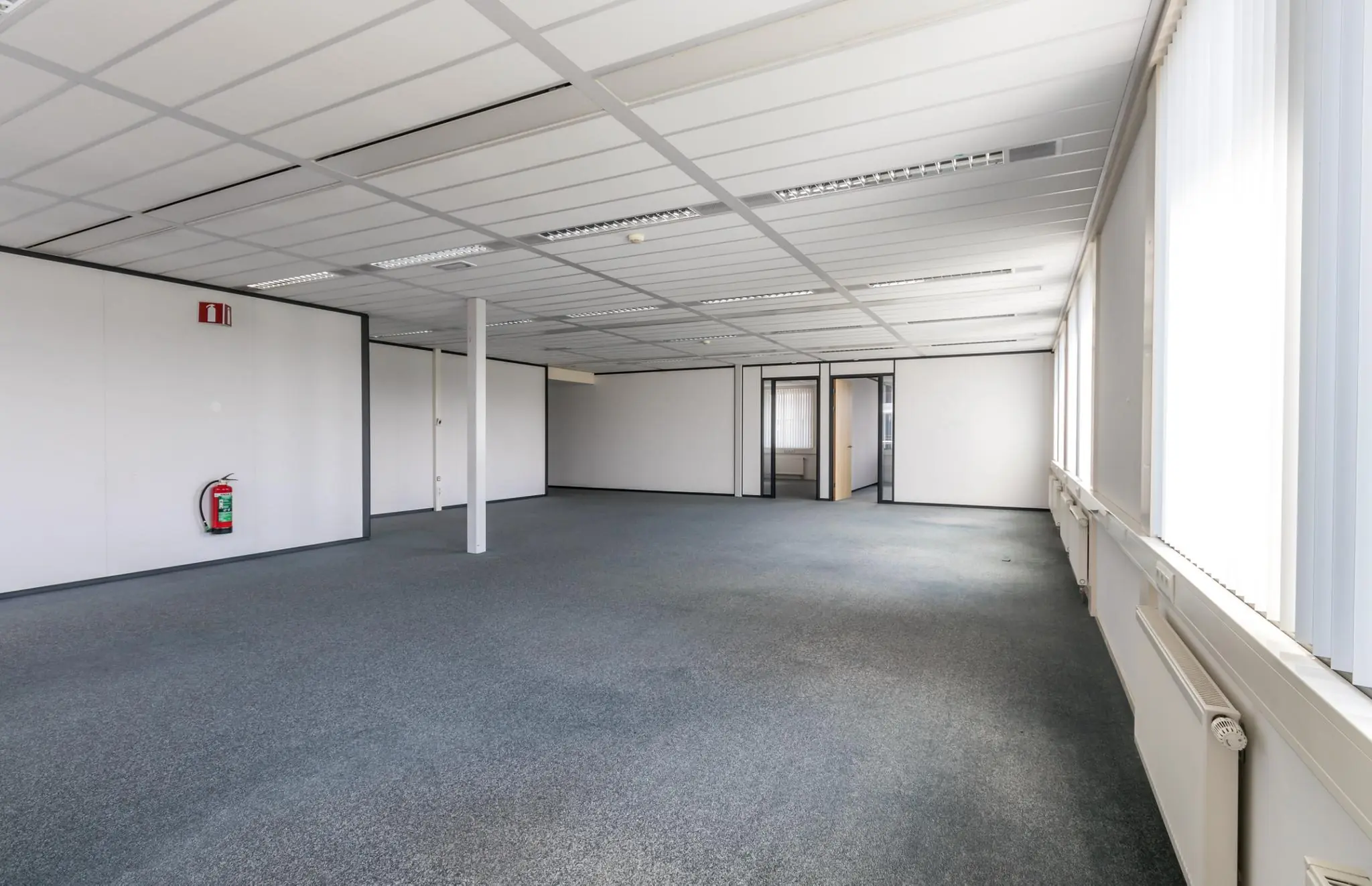 Lege kantoorruimte met systeemplafond, tapijtvloer en grote ramen met verticale lamellen.